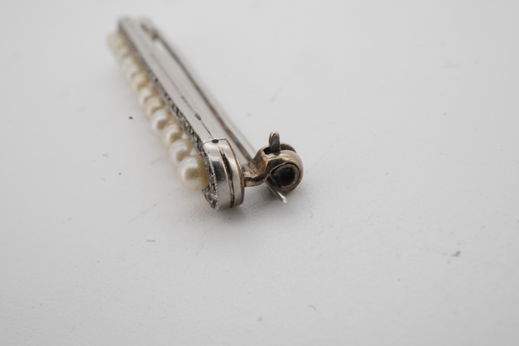 A Belle Epoque pearl and diamond brooch, early 20th century, of bar design, composed of a line of seed pearls within an oval border of rose-cut diamonds, mounted in platinum, length 3.9cm, French sponsor's mark, partial